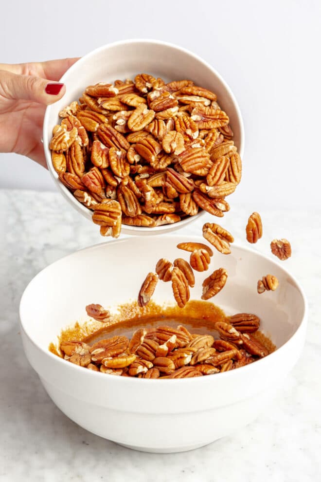 Hand pouring pecans from a white bowl into brown, wet mixture in a white bowl on a gray and white surface.
