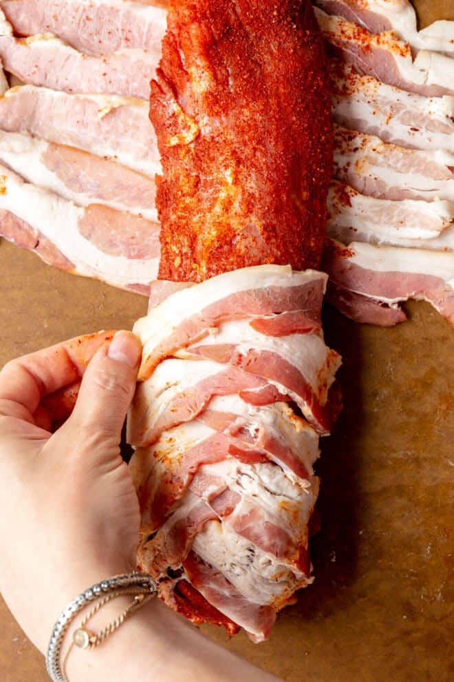 Hand folding over slices of bacon around seasoned pork tenderloin on a cutting board.