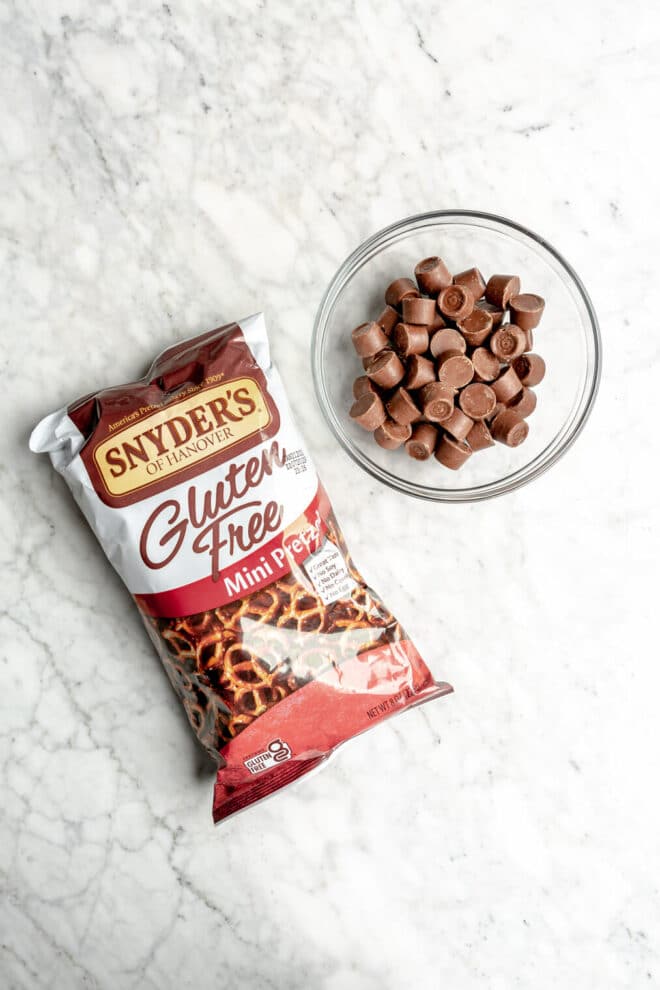 Bag of gluten free pretzels and a bowl of rolo candies in a glass bowl on a grey and white marble surface.