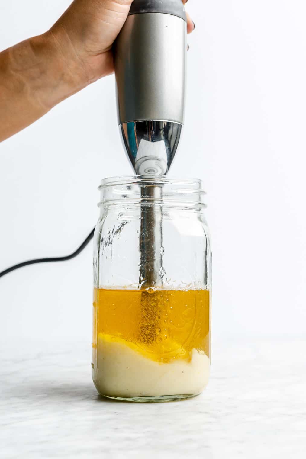 Hand holding immersion blender in a large mason jar as mayonnaise starts to form at the bottom and oil layered on top.