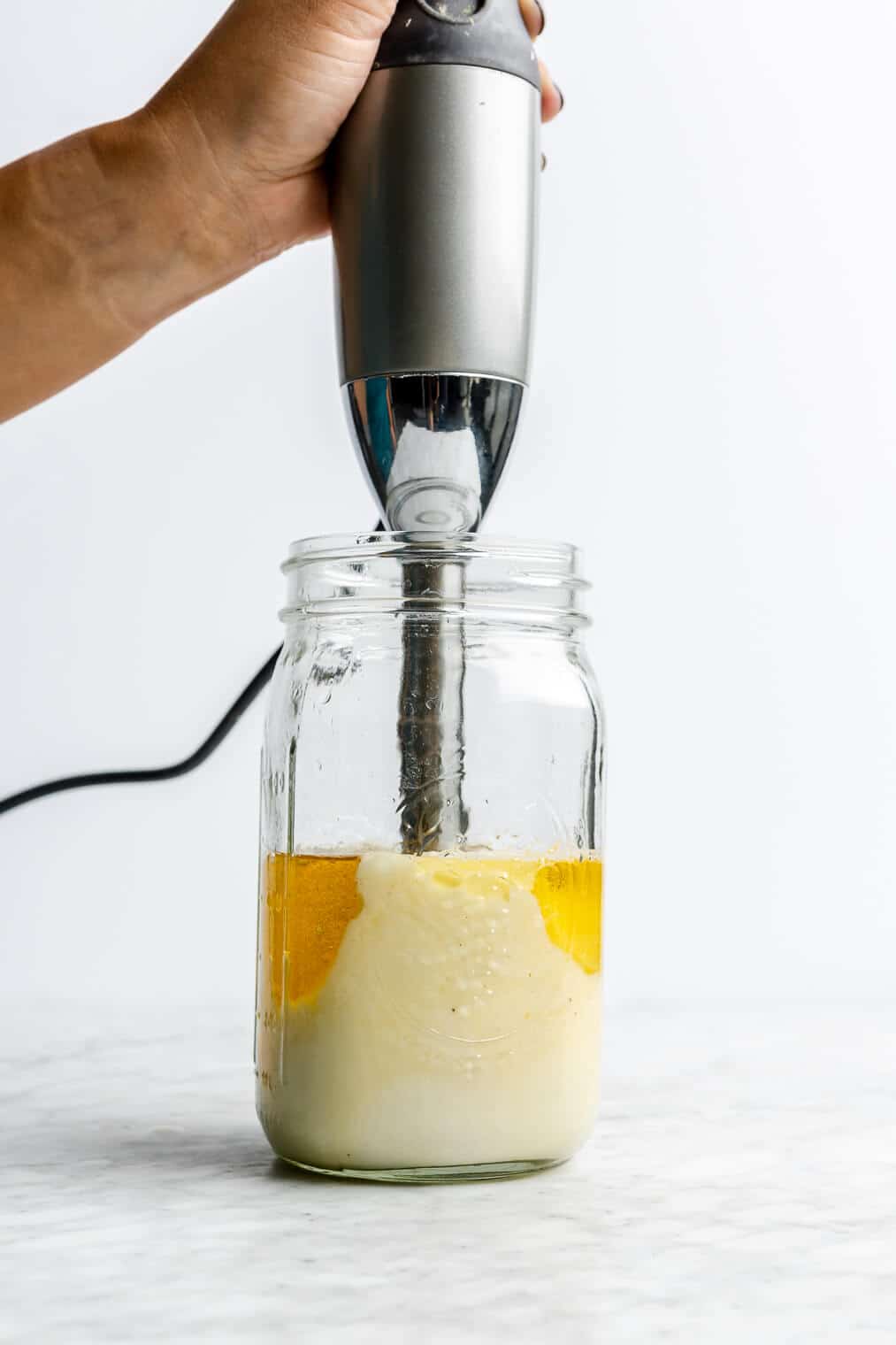 Hand holding immersion blender in a large mason jar blending homemade mayonnaise.