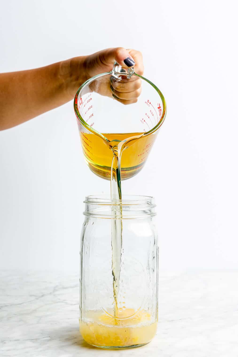 Hand pouring oil into large mason jar with cracked egg and lemon juice on a grey and white marble surface.