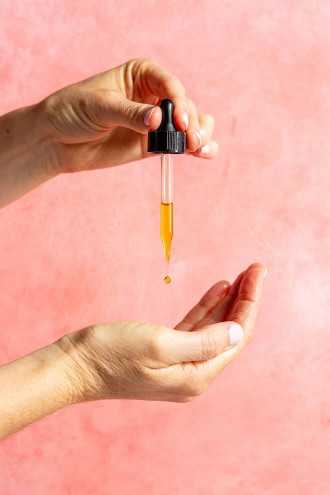Two hands, one holding a dropper and the other cupped below, with a drop of oil coming out of the tip of the dropper against a pink/blush background. 