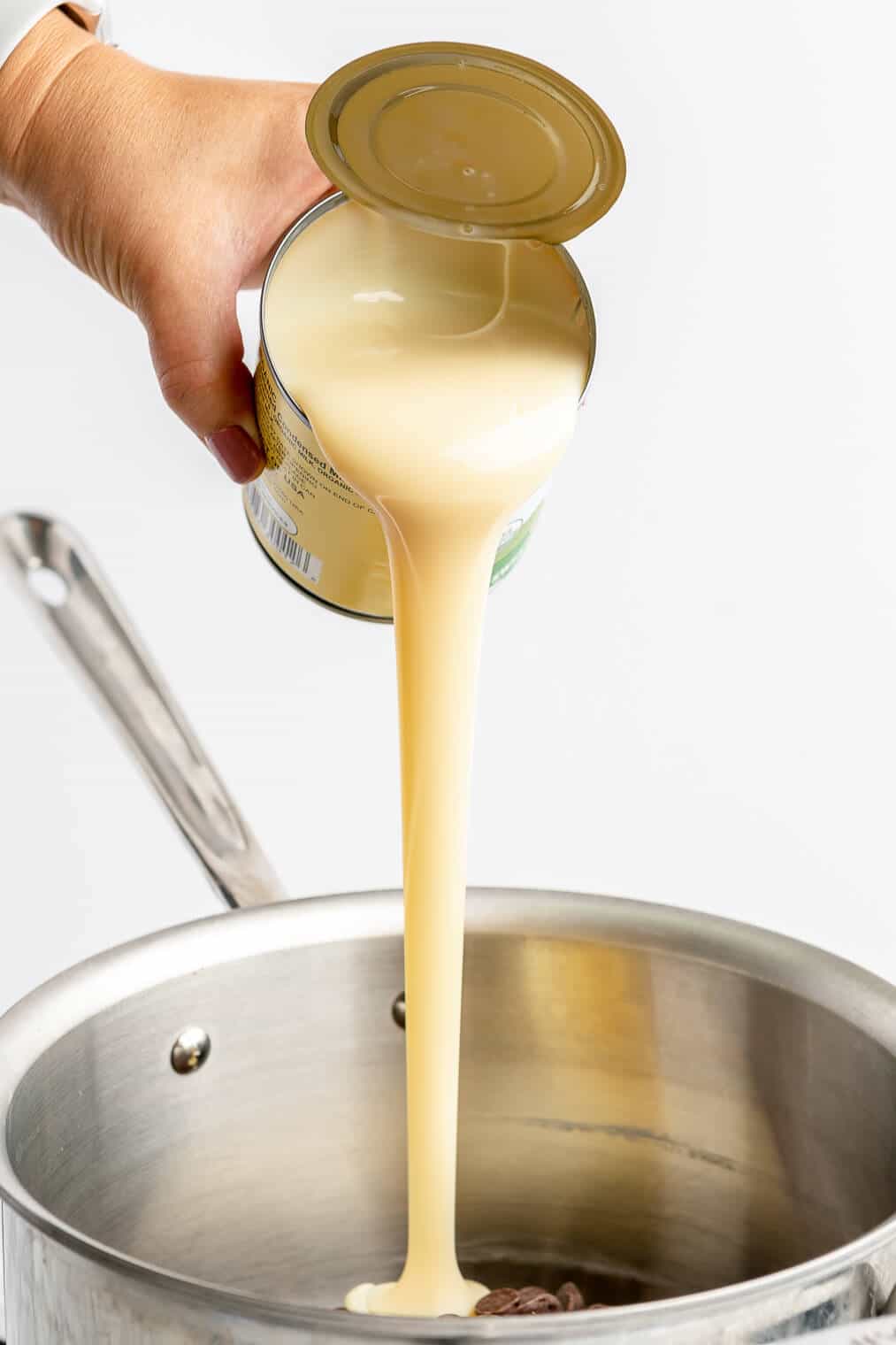 Hand pouring sweetened condensed milk into a stainless steel saucepan.