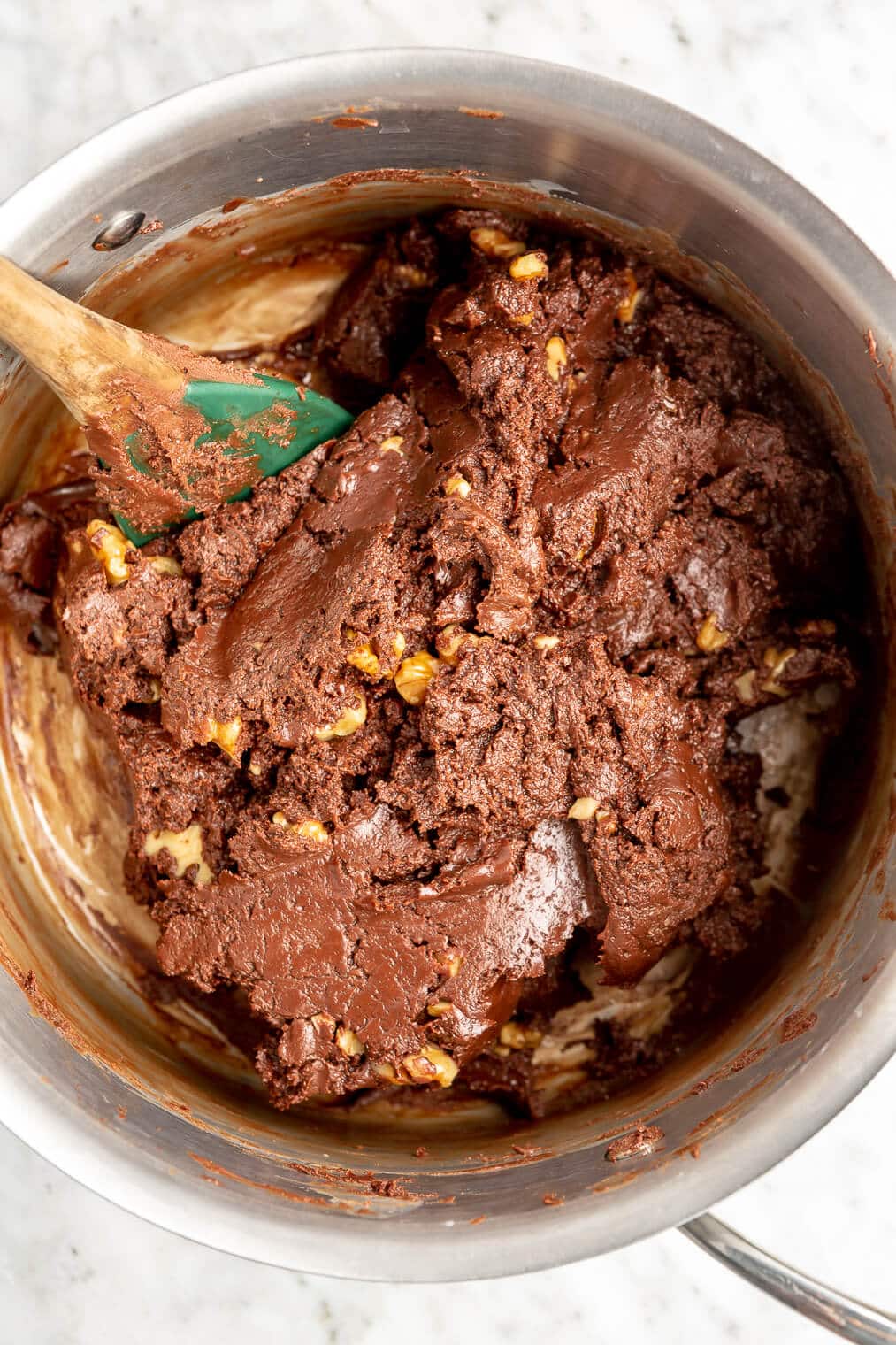 Spatula stirring melted chocolate, condensed milk, and walnuts in a stainless steel saucepan.