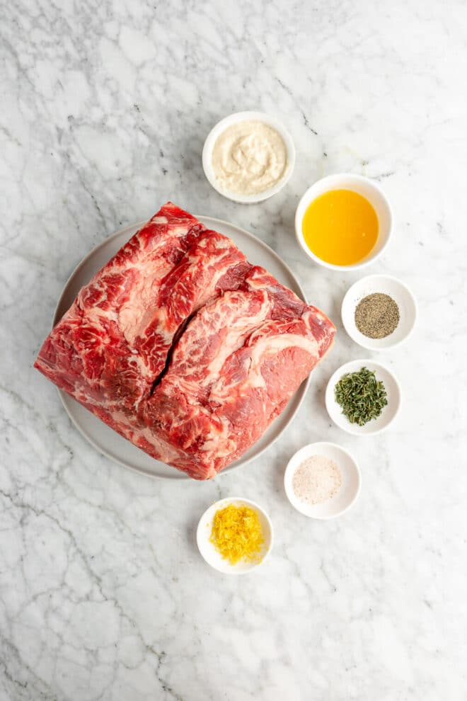 Prime rib roast on a plate surrounded by herb crust ingredients in small white bowls on a grey and white marble surface.