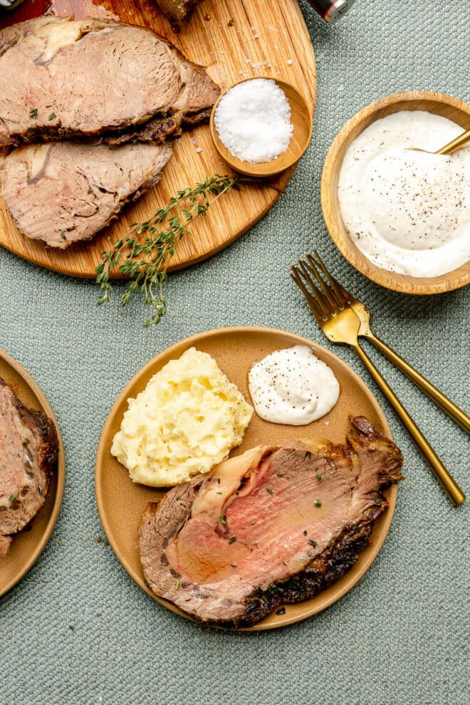 Top down photo of a plate of sliced prime rib served alongside mashed potatoes and a dollop of horseradish cream with gold silverware, a wooden bowl with horseradish sauce, and cutting board with more prime rib. 