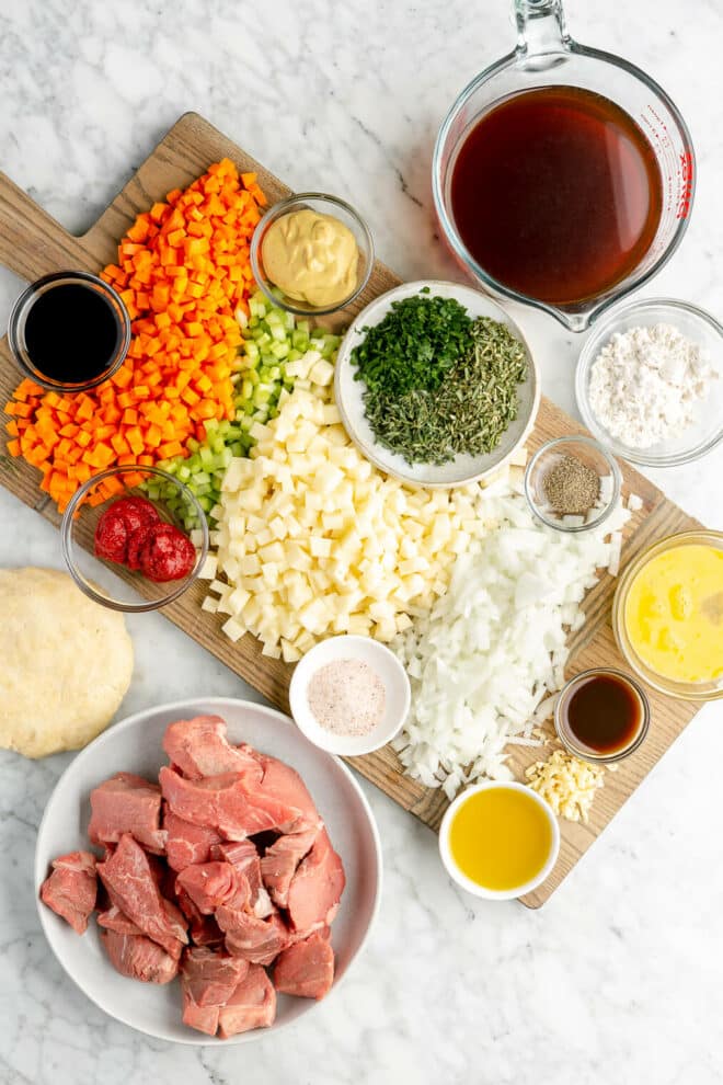 Top down photo of beef pot pie ingredients The vegetables are chopped on a rectangle cutting board. The remaining ingredients are in various glass and white bowls on a grey and white marble surface. 