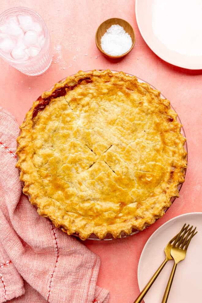 Top down view of beef pot pie on a pink surface. There are a couple of light pink plates to the top right and bottom left of the pot pie. The bottom plate has 2 forks on the plate. Also on the surface is a red and white striped towel, a glass of ice water, and a small wooden bowl with flaky sea salt.
