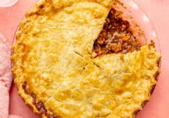 Top. down photo of beef pot pie with a flaky pie crust on top with one slice cut out. The pie dish is sitting on a pink surface and has a light pink plate with a gold fork to the top, a red and white striped linen to the bottom, and a light pink plate with a slice of pot pie served on it to the bottom right.