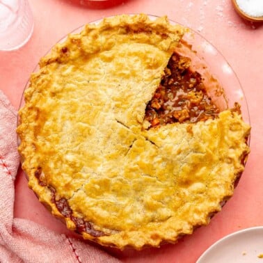 Top. down photo of beef pot pie with a flaky pie crust on top with one slice cut out. The pie dish is sitting on a pink surface and has a light pink plate with a gold fork to the top, a red and white striped linen to the bottom, and a light pink plate with a slice of pot pie served on it to the bottom right.