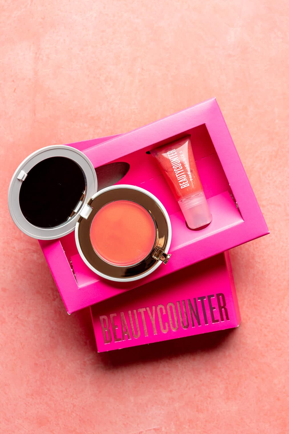 Top down view of Gloss and Glow duo. The cream blush is open next to a lip jelly. Both are sitting on top of a fuchsia box on a pink blush surface.