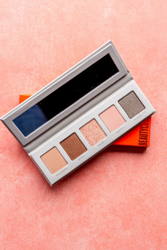 Beautycounter essentials eye palette. The palette contains 5 different eye shadows and a rectangle mirror. It is sitting on top of a red, rectangle box on a blush pink surface. 