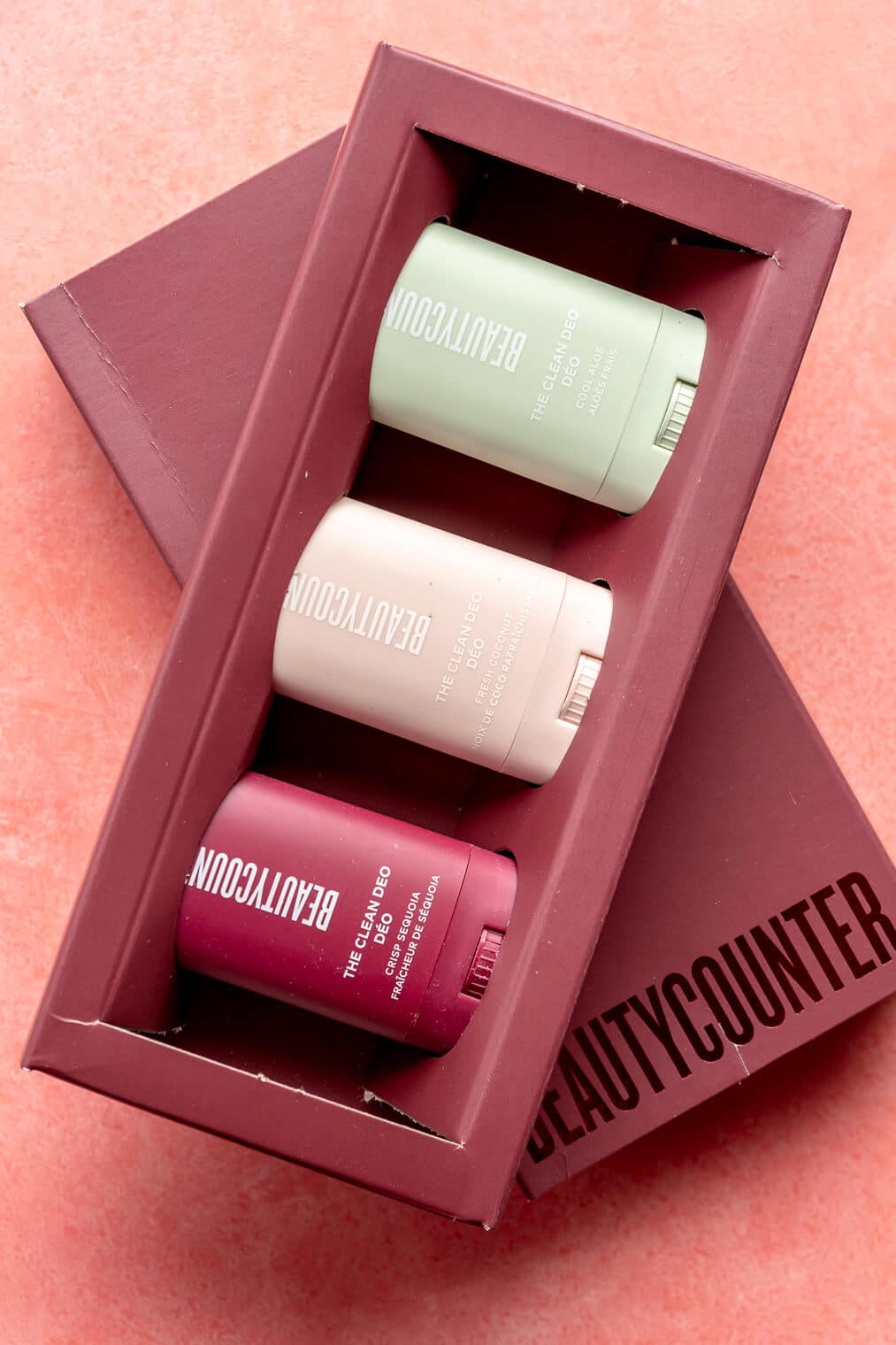 Three miniature deodorants in a rectangle maroon box sitting on a pink blush surface.