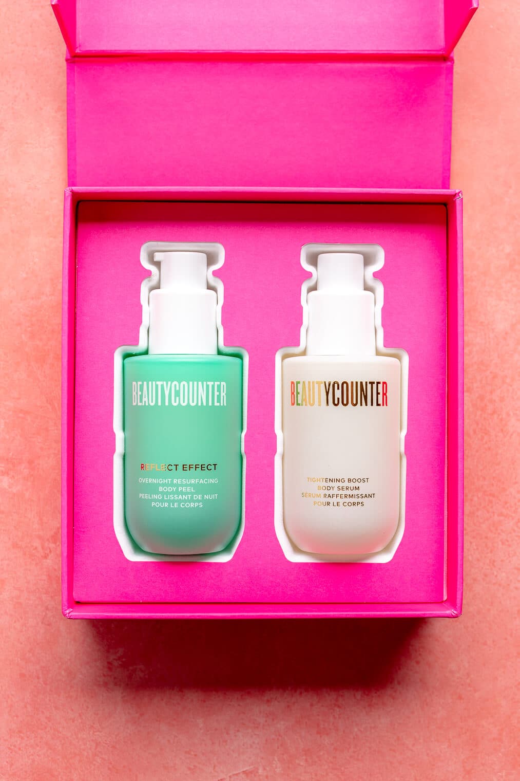 Two bottles, one is turquoise, one is white, nestled in a fuchsia box on a pink blush surface.