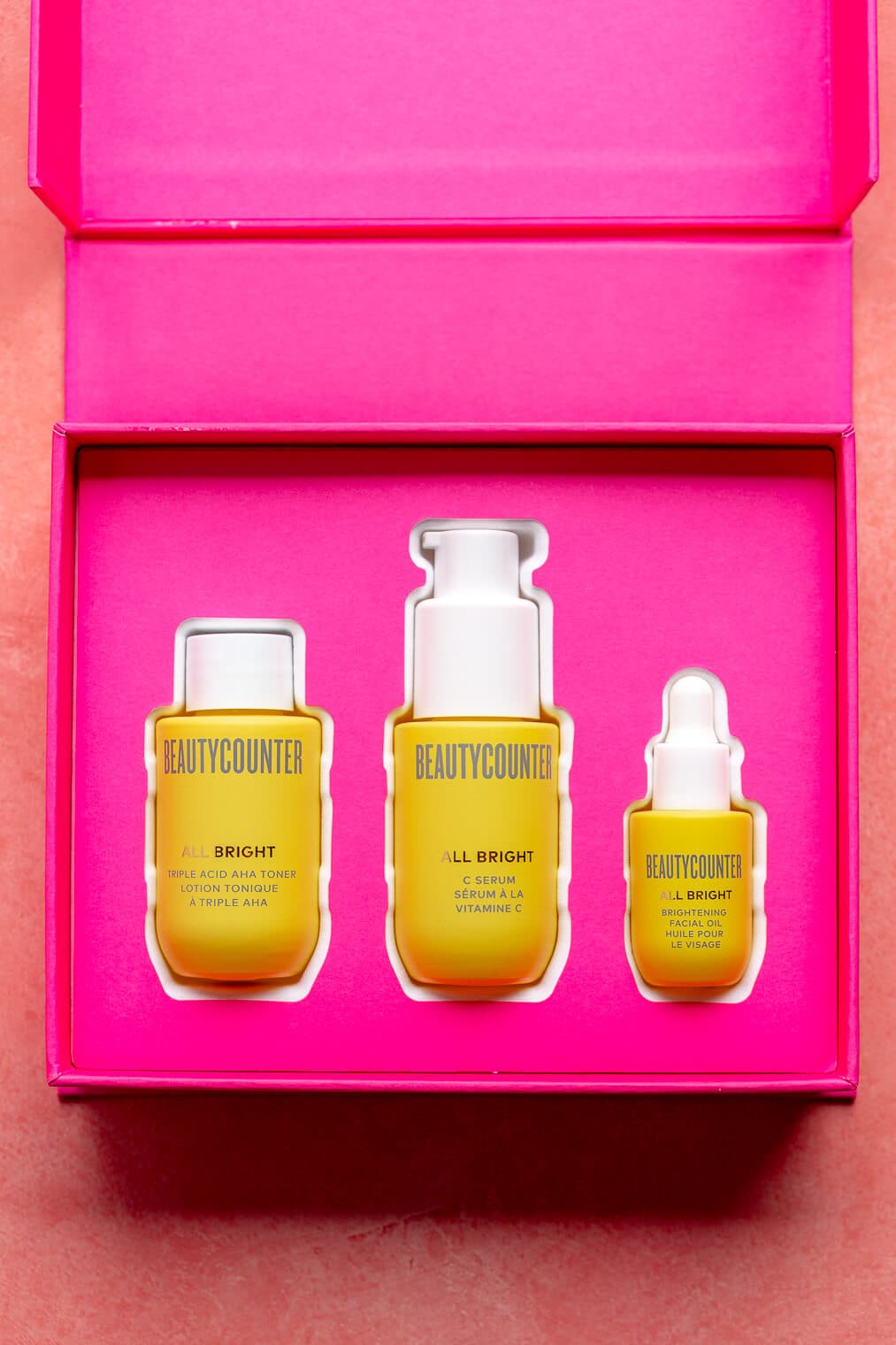 Mini AHA toner, C serum, and brightening oil in yellow bottles nestled in a fuchsia box.