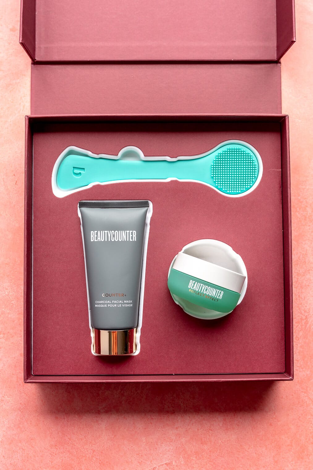 A turquoise silicone skin care tool is nestled in the top of the box. Below it are a charcoal tube and a small, round, turquoise container containing face masks. All are sitting in a maroon box on a pink blush surface.