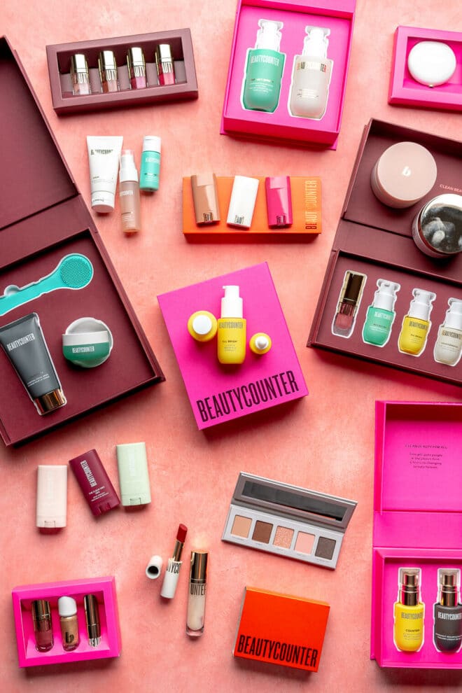 Top down view of Beautycounter limited edition sets. All of the boxes and sets are open so that you can see the products inside. They contain a variety of beauty and skincare items, including makeup, lotions, masks, deodorants, and lip glosses. The packages are fuchsia, maroon, and red in color. 