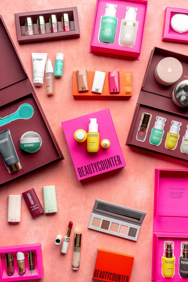 Top down view of Beautycounter limited edition sets. All of the boxes and sets are open so that you can see the products inside. They contain a variety of beauty and skincare items, including makeup, lotions, masks, deodorants, and lip glosses. The packages are fuchsia, maroon, and red in color.