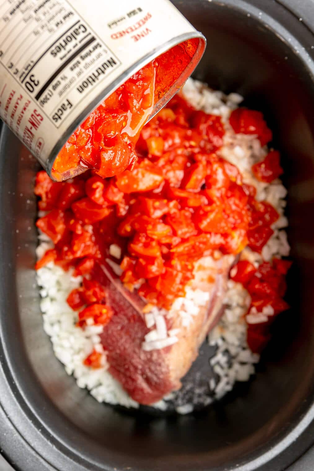 Canned diced tomatoes being poured into a crockpot with beef roast and onions.