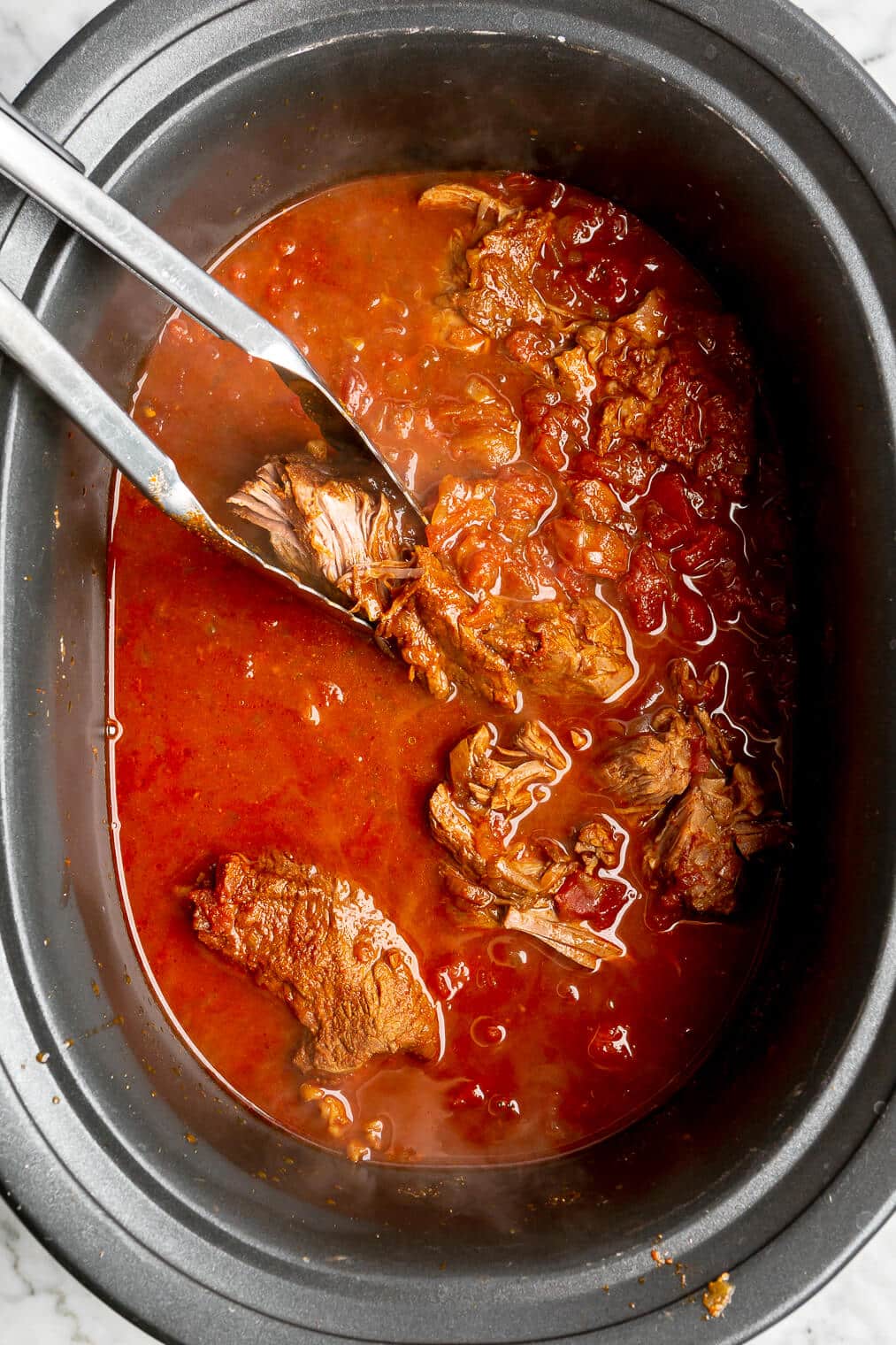 Tongs shredding cooked beef in a crockpot in a red liquid.