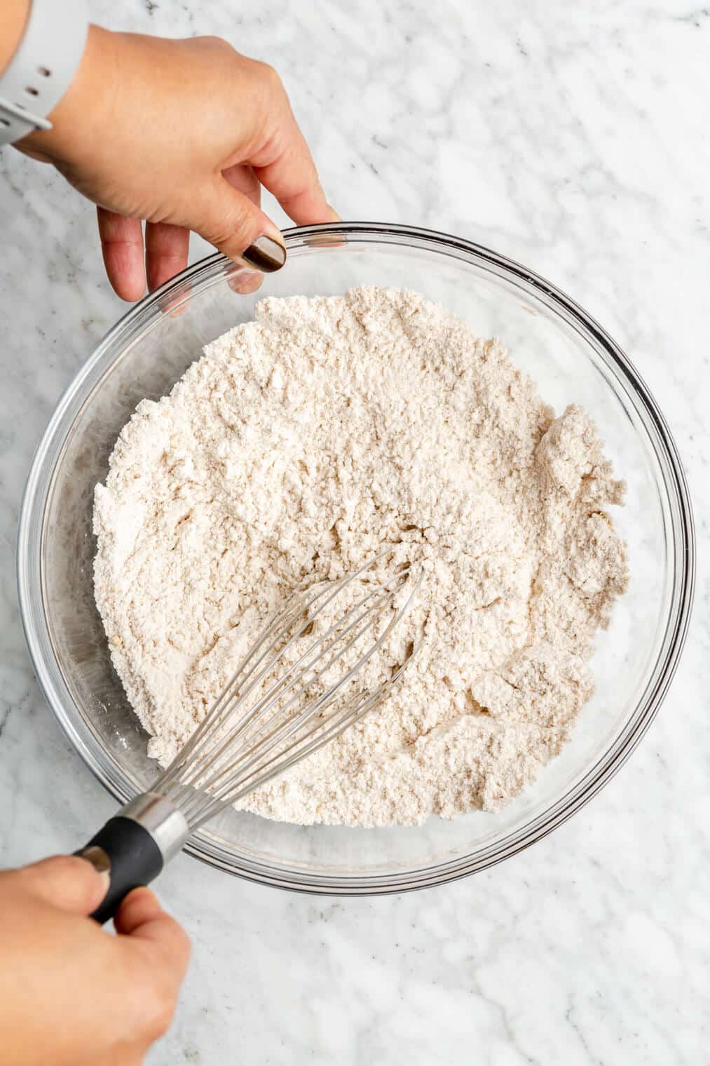 Two hands holding a glass bowl with a whisk mixing together dry flour ingredients in the bowl.