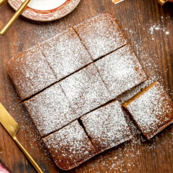 Classic gingerbread sprinkled with powdered sugar cut into nine square slices on a dark wooden cutting board. There is a dark red, rimmed plate in the top corner with gold silverware and two cups of tea in decorated tea cups.