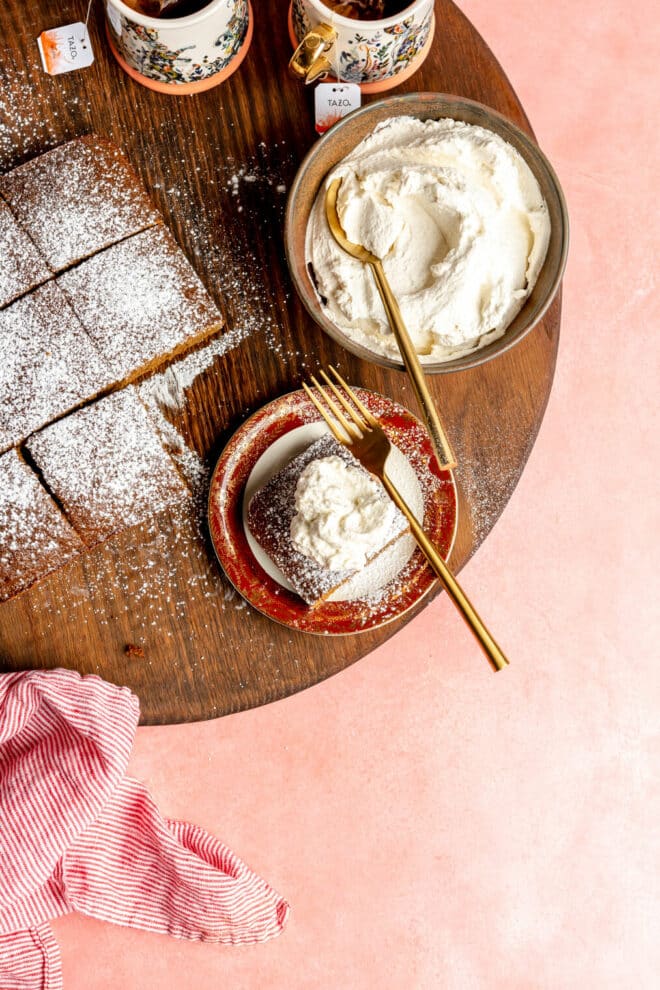 A corner slice of classic gingerbread served on a maroon rimmed, white plate with a gold fork sitting on a round, wooden cutting board. The plate is next to the gingerbread loaf and a bowl of whipped cream with two cups of tea. 