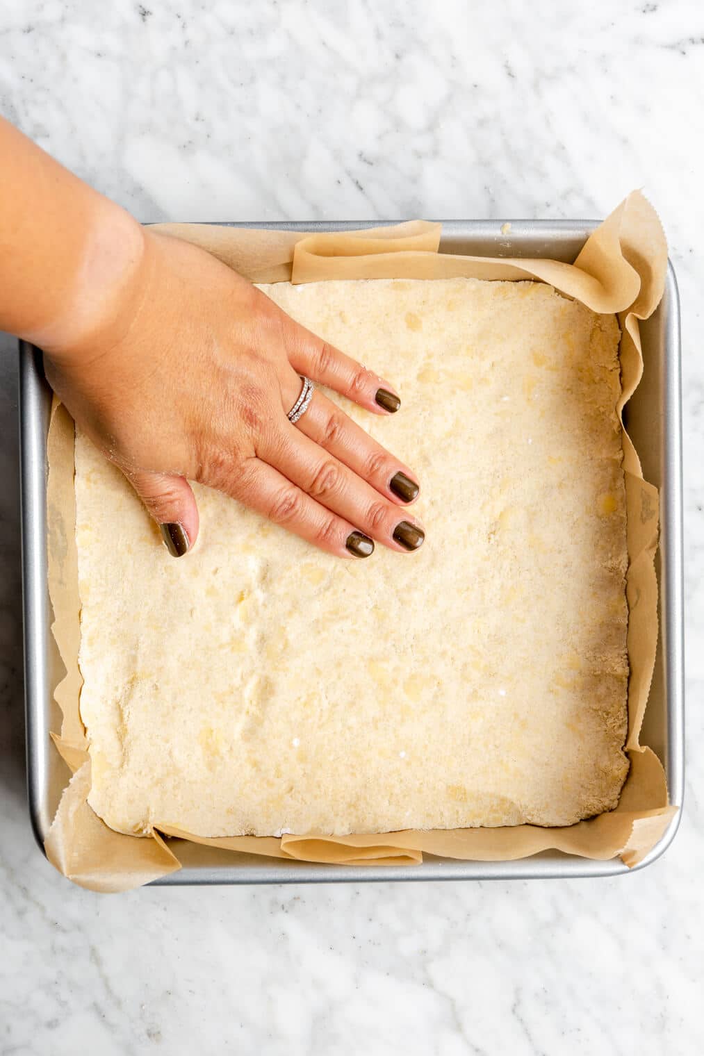 Hand pressing shortbread into an 8x8 baking pan lined with parchment paper.