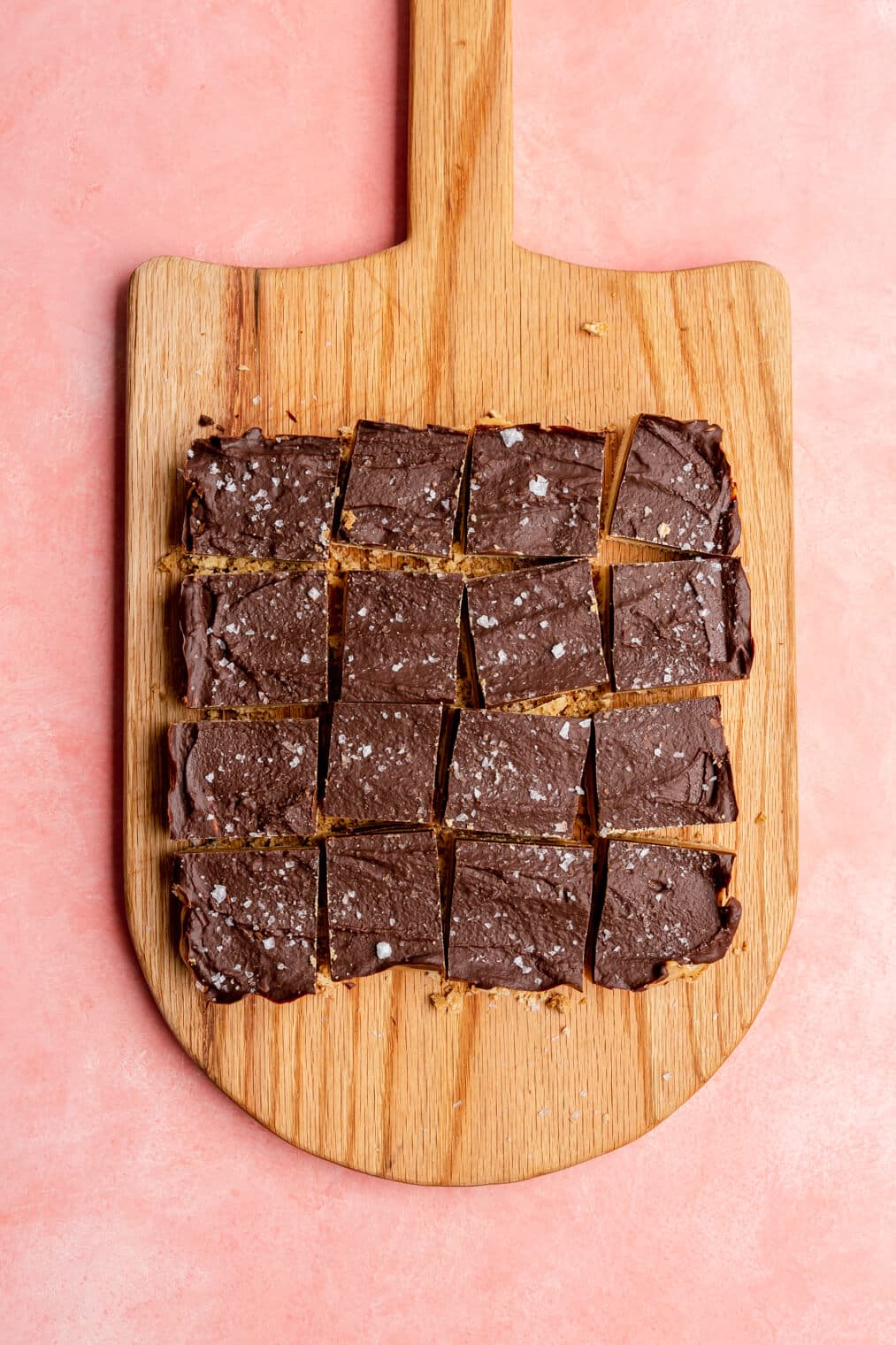 Millionaire shortbread sliced into square pieces on a wooden cutting board on a pink surface.