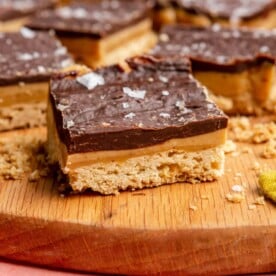 Side view of millionaire shortbread on a wooden cutting board. You can see the three layers of shortbread, caramel, and chocolate ganache that is topped with a flaky sea sat.
