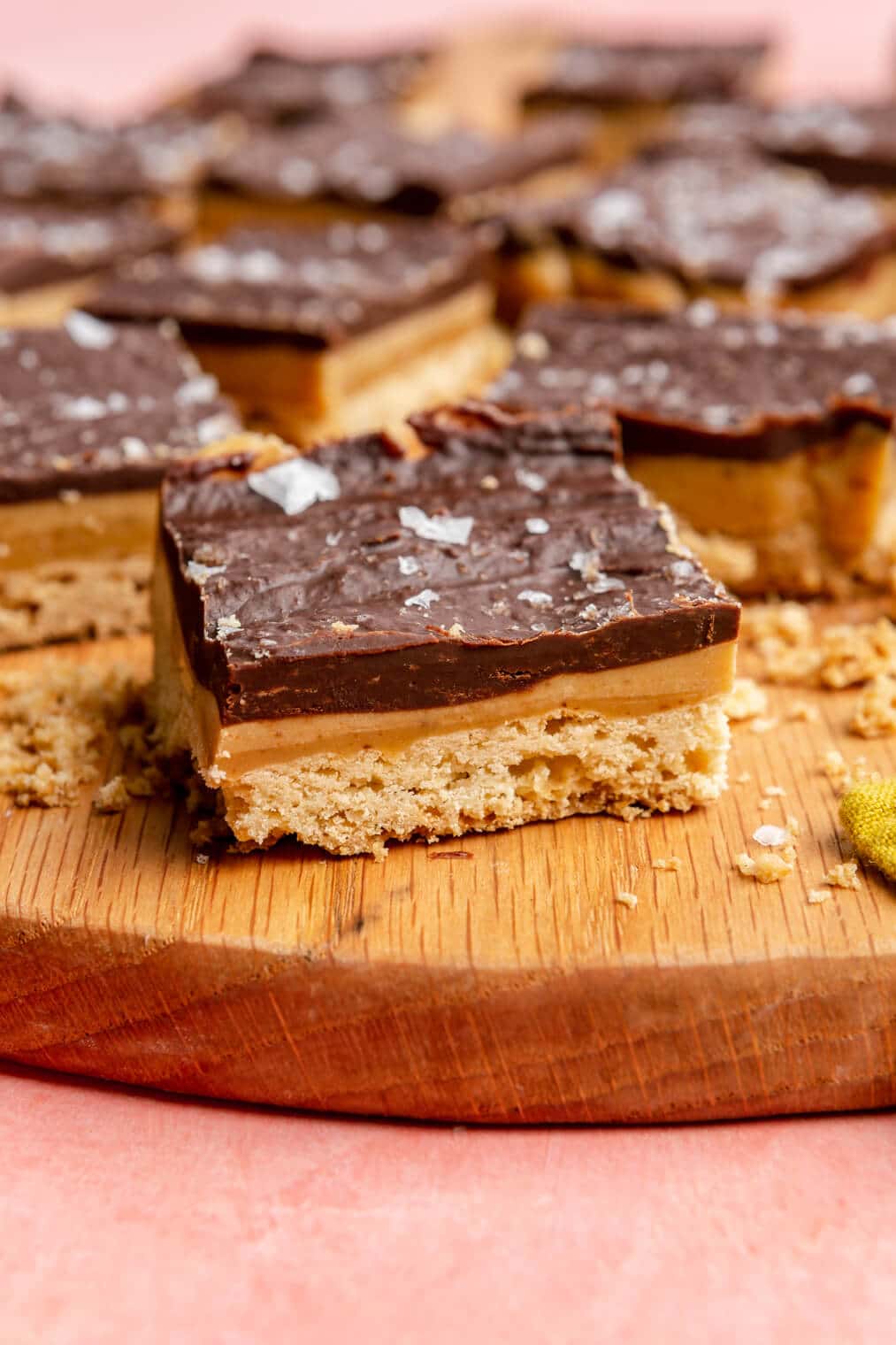 Side view of millionaire shortbread on a wooden cutting board. You can see the three layers of shortbread, caramel, and chocolate ganache that is topped with a flaky sea sat.