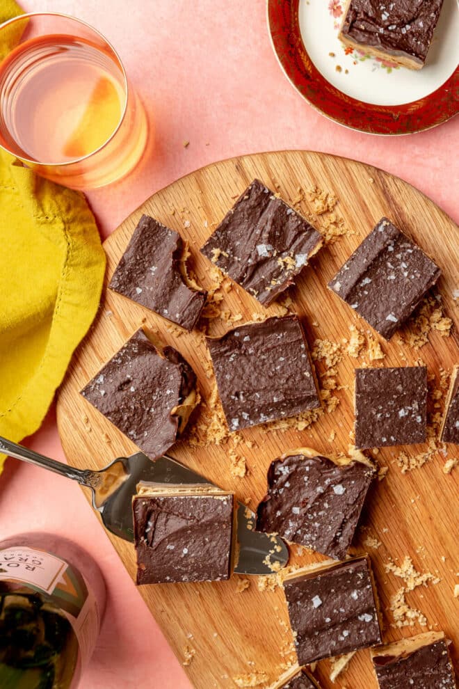 Pieces of millionaire shortbread on a wooden cutting board on a pink/blush surface. One piece is sitting on a silver server. There is a glass of wine in the background with a mustard yellow linen. There is a maroon rimmed plate with a piece of millionaire shortbread on the plate. 