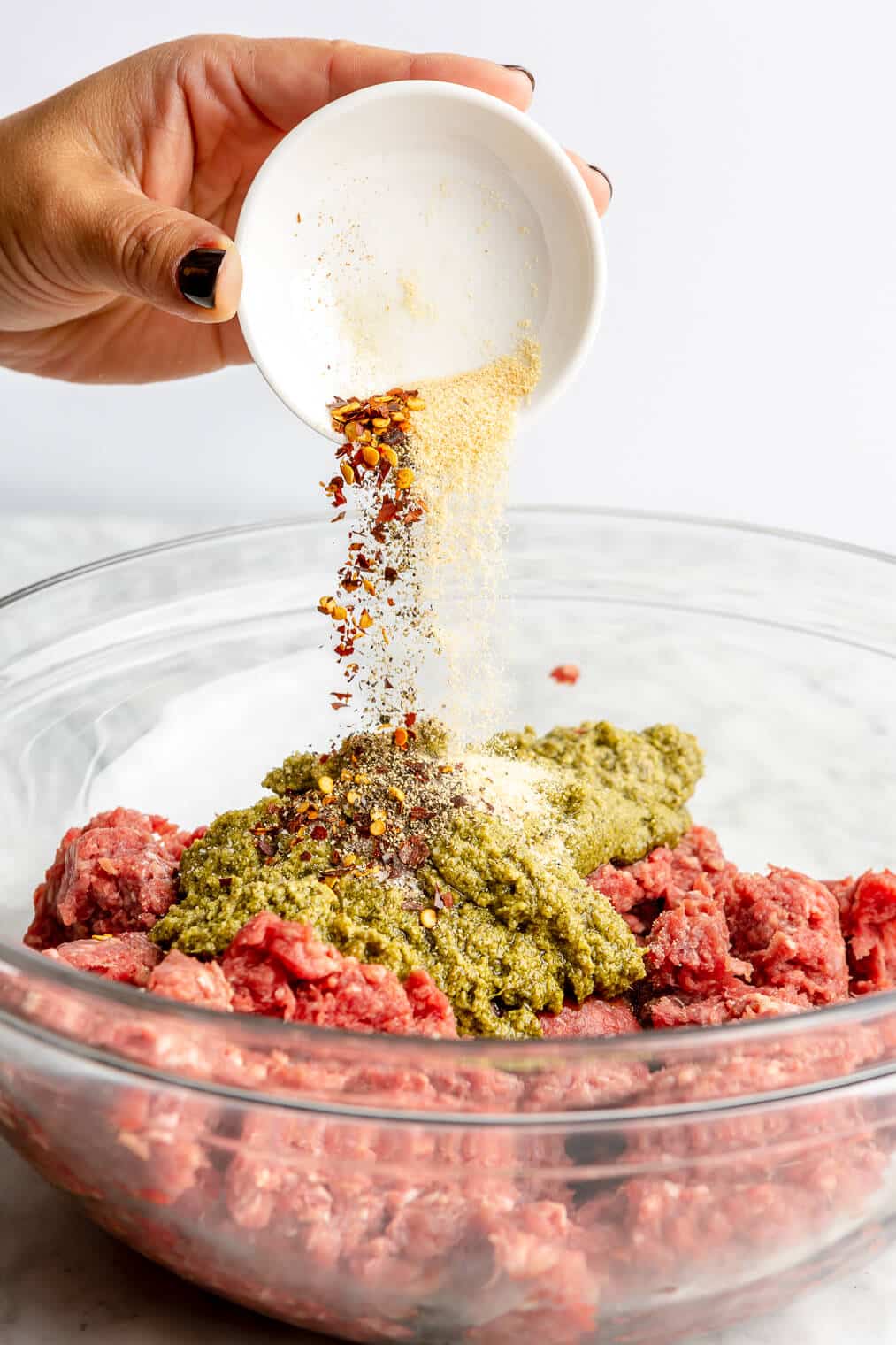Hand holding small, white bowl pouring spices into a ground beef mixture topped with pesto.