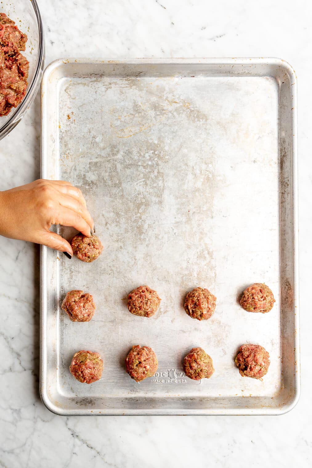 Hand placing meatballs in rows of 4 on a baking sheet.