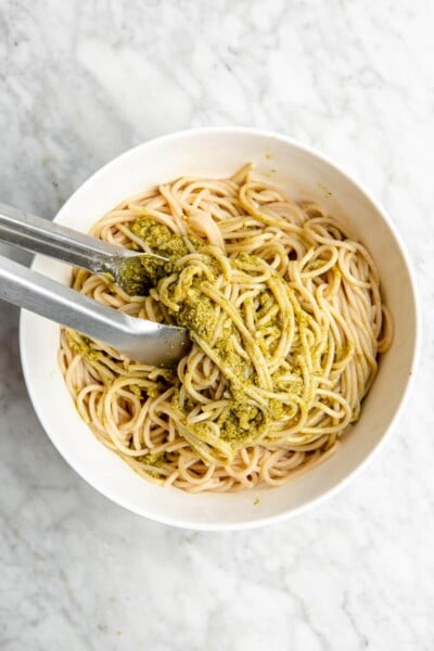 Tongs tossing spaghetti noodles with pesto.