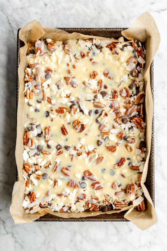 Layers of chocolate and white chocolate chips, coconut flakes, and pecans covered with sweetened condensed milk in a 9x13 pan lined with parchment paper.