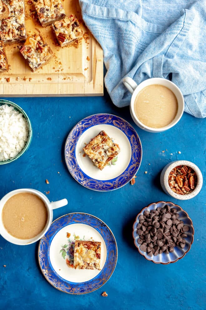 Top down view of a blue table top with a cutting board with magic bars sliced, a couple of coffee mugs, two blue rimmed plates with a magic bar plated on the plates, a bowl of chocolate chips, a small serving dish with pecans, and another serving dish with coconut flakes. 