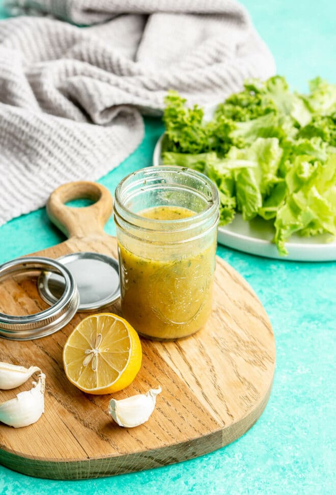 Jar of apple cider vinaigrette on a wooden cutting board. There is half a lemon and garlic cloves on the cutting board a well and a plate of lettuce in the background. All are sitting on a teal surface.