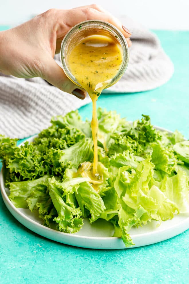 Hand pouring salad dressing onto a plate of green leaf lettuce. The plate is sitting on a bright teal surface, and there is a grey linen draped in the background.