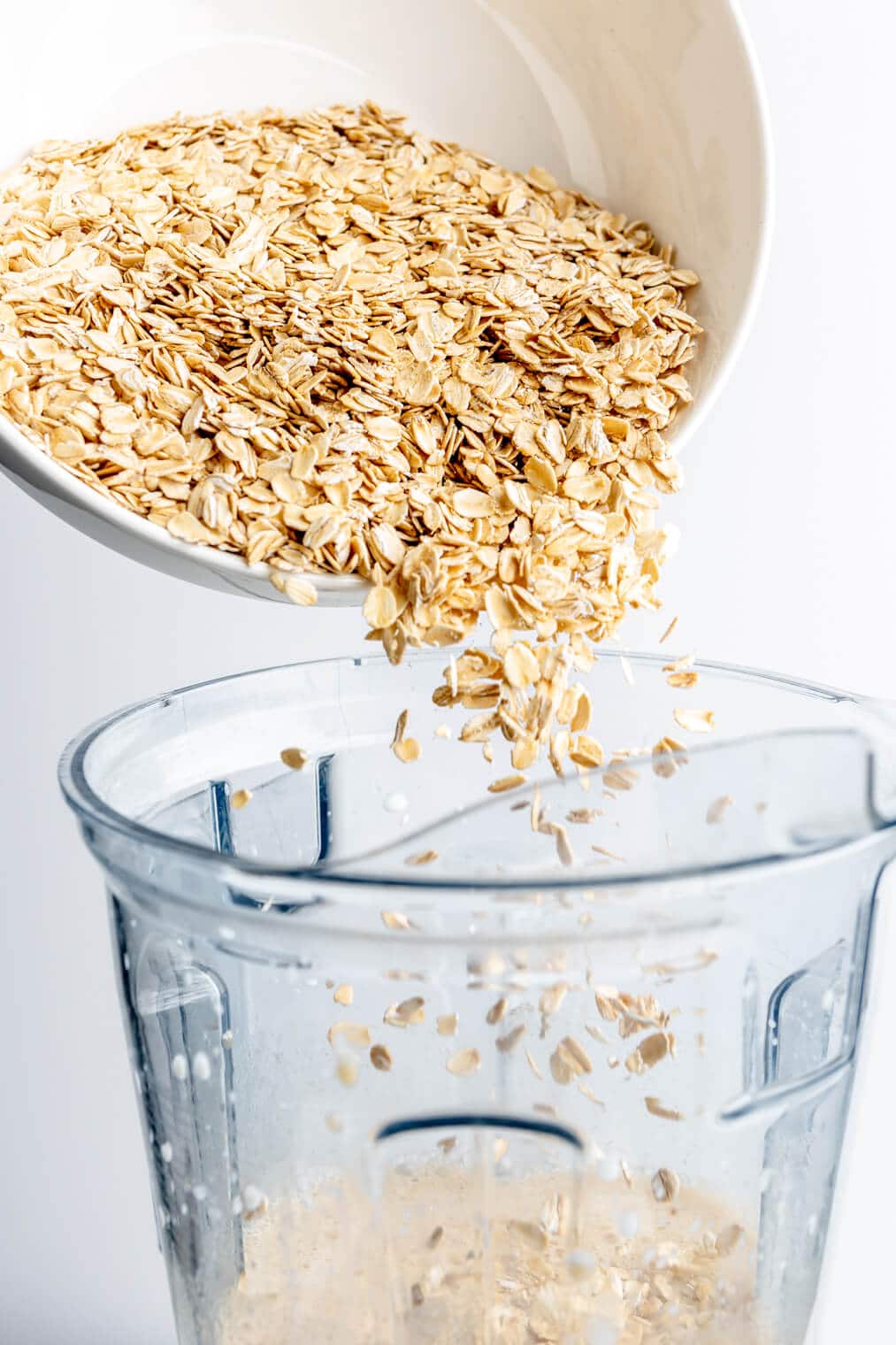 Oats being poured into a blender pitcher from a white bowl.
