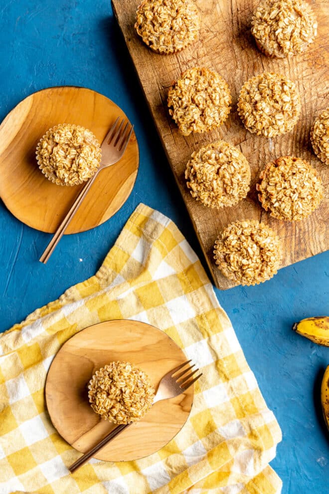 Top down view of baked oatmeal cups on a blue surface with a yellow and white checkered linen. There are some cups that are on a wooden cutting board and there are individually plated muffins on round, wooden plates with gold forks.