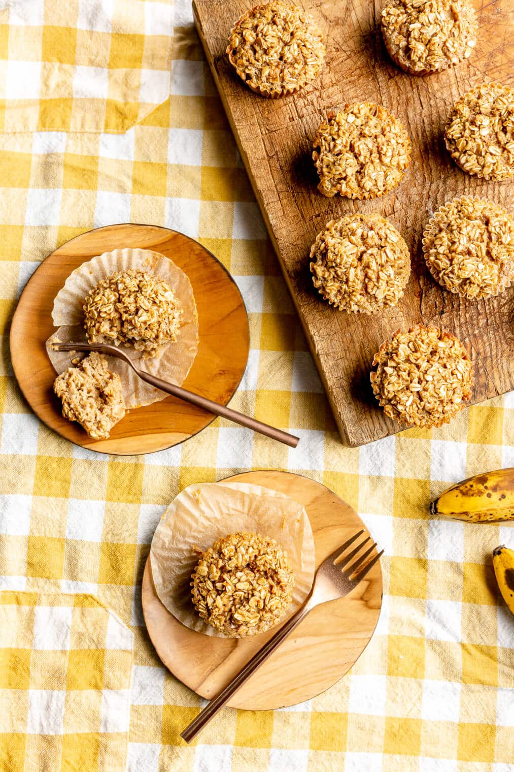 Top down view of baked oatmeal cups on a yellow and white checkered linen. There are some cups that are on a wooden cutting board and there are individually plated muffins on round, wooden plates with gold forks.