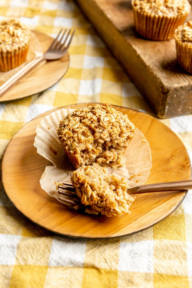 A baked oatmeal cup on a round, wooden plate. There is a piece sliced out of the muffin and sitting on a gold fork. The muffin is sitting on a cupcake liner. There is a wooden cutting board behind the muffins and another plate with a muffin in the background. Everything is on a surface covered with a yellow and white checkered linen.