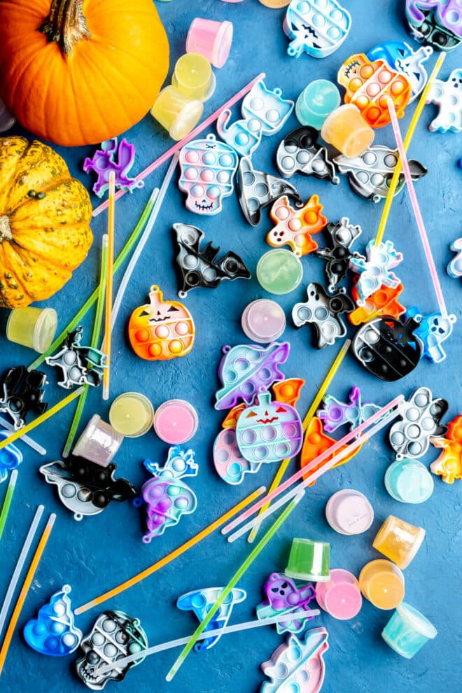 Variety of keychain pop beads, small containers of slime, glow sticks, and a couple of small pumpkins all on a blue surface.