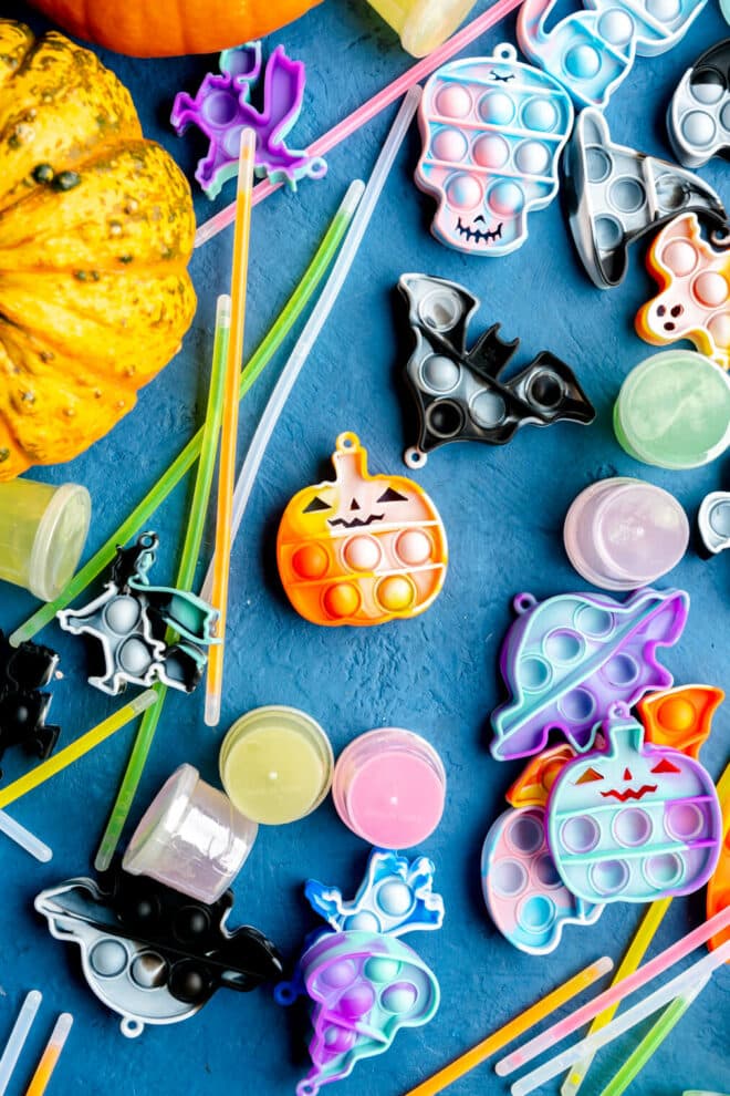 Variety of keychain pop beads, small containers of slime, glow sticks, and a couple of small pumpkins all on a blue surface.