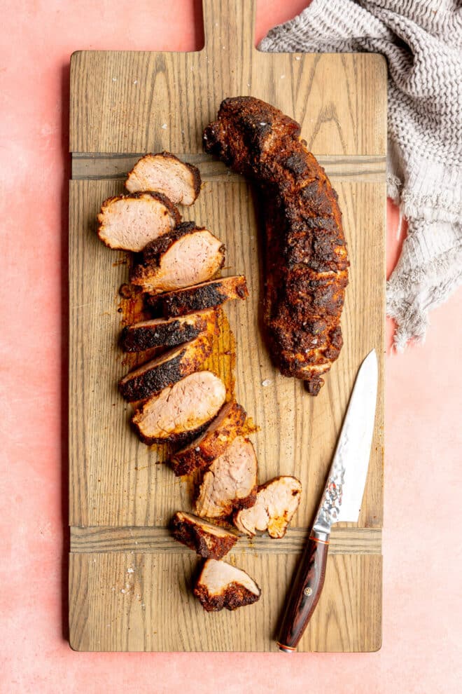 A rectangle, wooden cutting board with two roasted pork tenderloins. One is sliced into medallions, and the other is whole. Both have a dark chili rub on the exterior. 