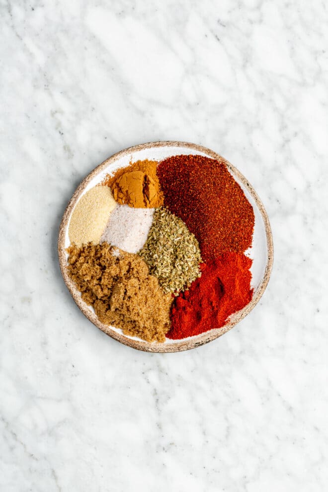 Top down view of dry rub ingredients on an imperfect round dish. The dish is sitting on a grey and white marble surface.