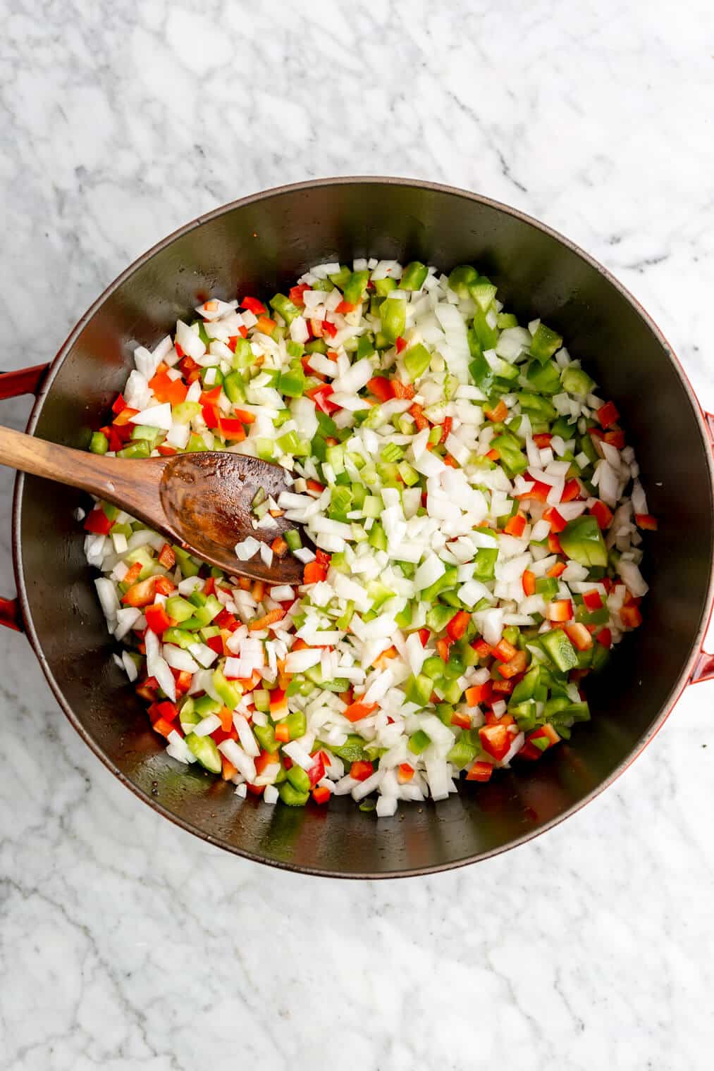 Diced onion, bell peppers, garlic, and celery being sautéed in a large dutch oven with a wooden spoon.
