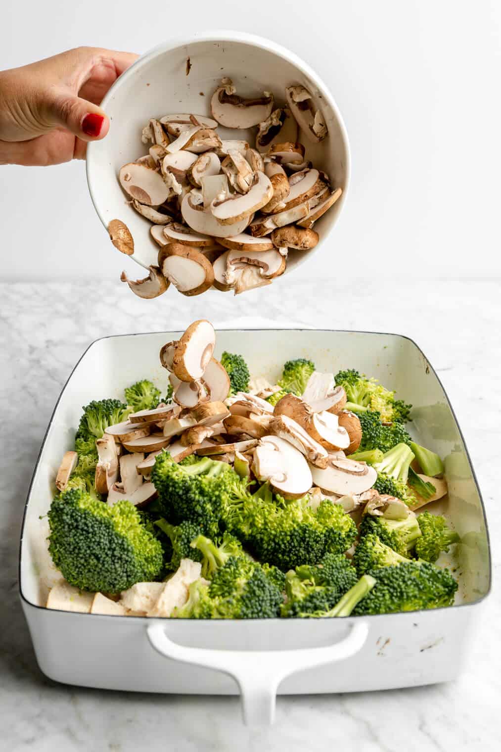 Hand holding a white bowl pouring sliced mushrooms over broccoli and chicken in a casserole dish.